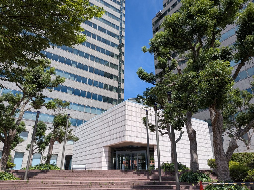 大きな並木の参道の奥に、石張りのエントランスと高層タワーが見える横浜ビジネスパーク
