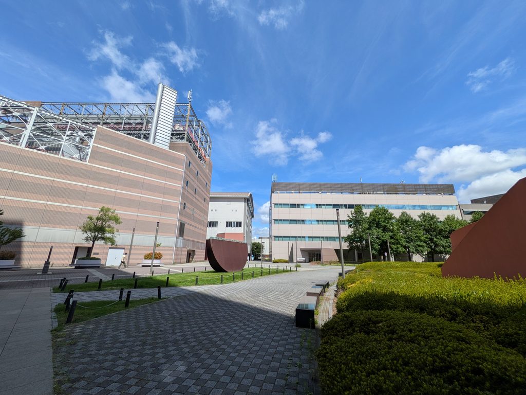 ブロック舗装の小道を一人が歩き、芝生に錆色の彫刻、背後にオフィス棟が見える横浜ビジネスパーク