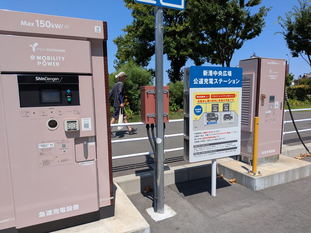 急速充電器と公道充電ステーションの看板のすぐ脇を、買い物袋を提げた男性が歩いている