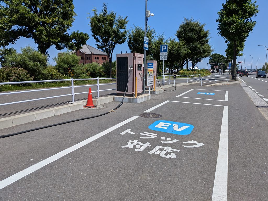 青空の下、赤レンガ倉庫を背景に、急速充電器と路面の大きな「EVトラック対応」表示が並ぶ新港中央広場の公道充電ステーション