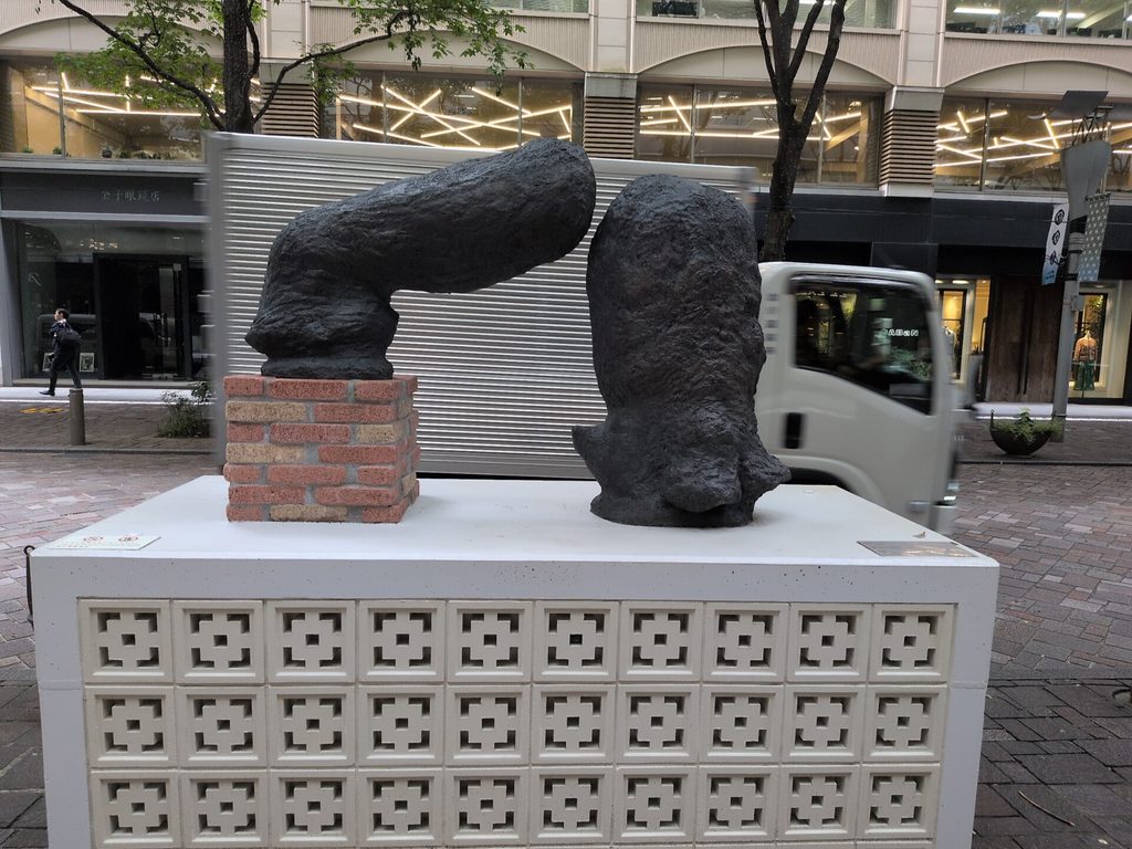 白い台座の上に、二つの黒い抽象彫刻が並ぶ。左は赤茶色のれんが台に載り、背景に店舗と走行中の車両が見える。