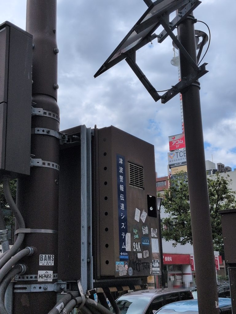 屋外の柱に取り付けられた箱形の設備。前面に「津波警報伝達システム」と表示され、柱の上部に太陽電池パネルとみられる板状の部材がある