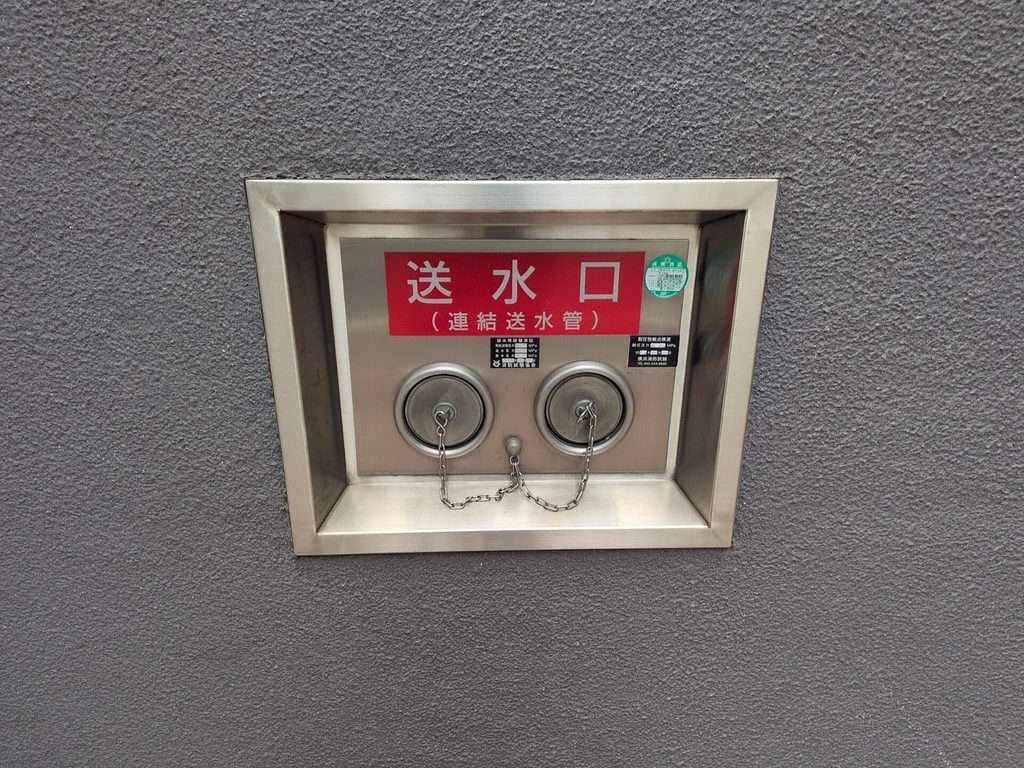 建物の外壁に埋め込まれた2つの接続口。「送水口(連結送水管)」と表示され、キャップと鎖が付いている。右上に緑色の円形ラベルが貼られている