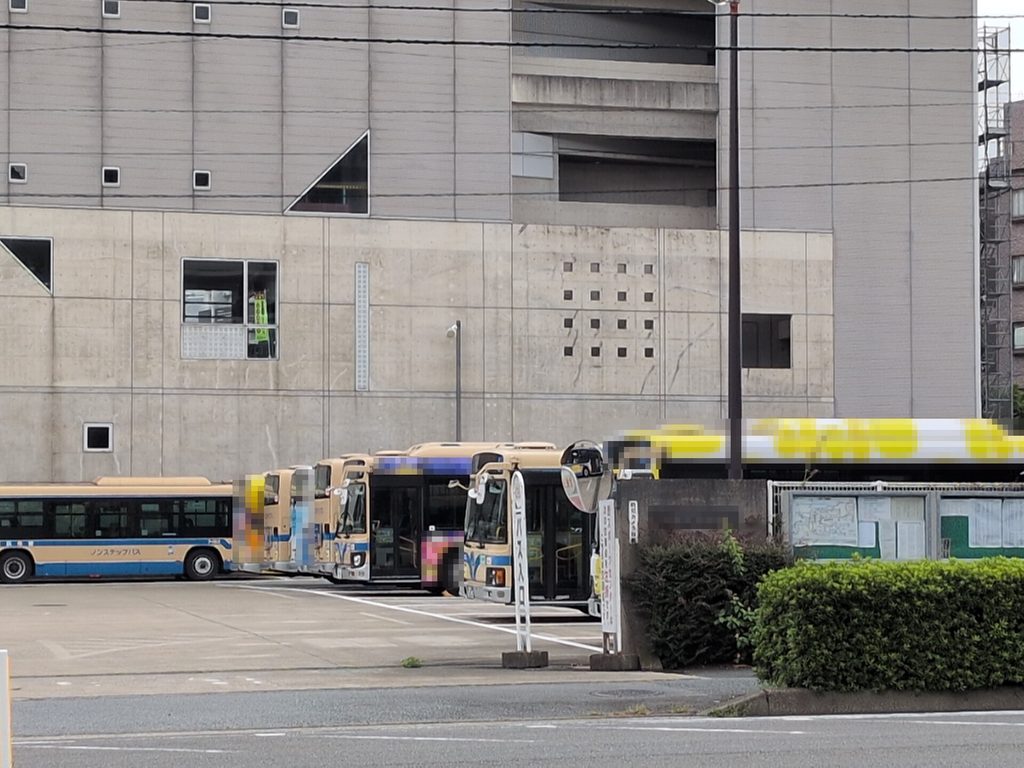 灰色の大きな建物の前の舗装された空間に、複数のバスが並んで停まっている。手前には案内板と植え込みがある