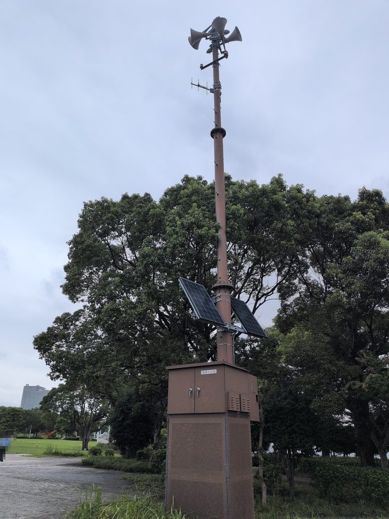 公園の樹木の前に立つ茶色の高い柱。上部に複数方向を向いたホーン形のスピーカーと横棒状のアンテナのような部材、中ほどに2枚の太陽電池パネル、足元に箱形の筐体がある