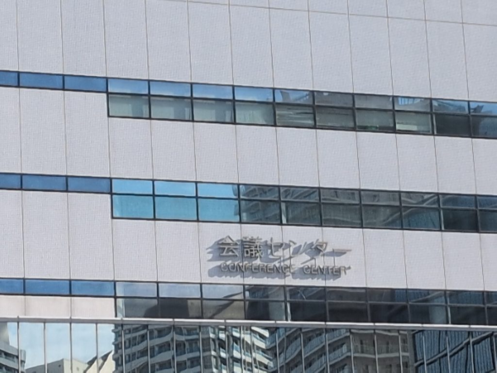 白色系の外壁に、日本語の「会議センター」と英語の「CONFERENCE CENTER」の立体表示がある。窓には周辺の建物が映り込んでいる