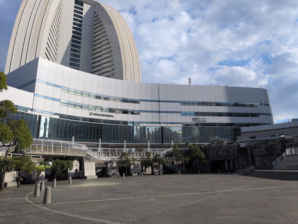 広場の向こうに、白色系の低層の建物が横に広がり、外壁に「会議センター」の表示がある。その背後に曲面を含む高層の建物が隣接して見え、手前には階段と歩行者用の通路、樹木が見える
